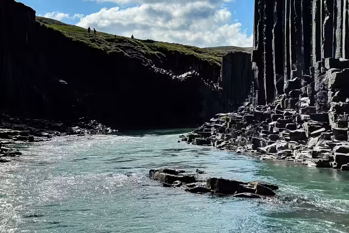Towering dark basalt columns at Studlagil Canyon with two hikers silhouetted on green hillside under blue sky