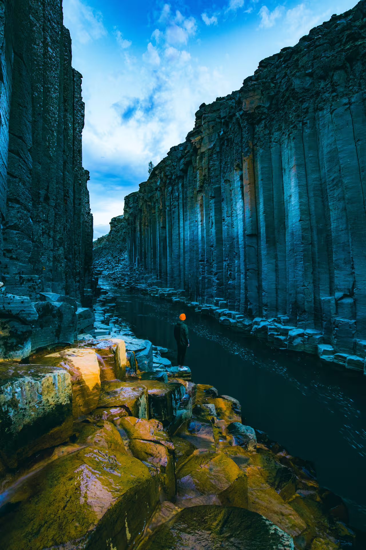 Hiker in orange hat standing beside dark basalt column walls of Studlagil Canyon with teal river below