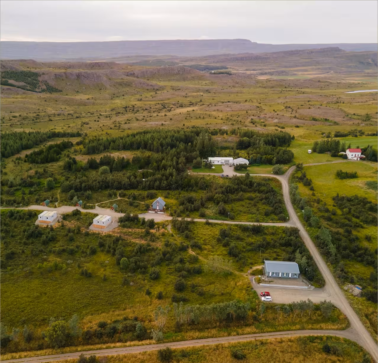 Aerial view of rural Icelandic landscape with scattered buildings, lush greenery, and dramatic plateau cliffs in background