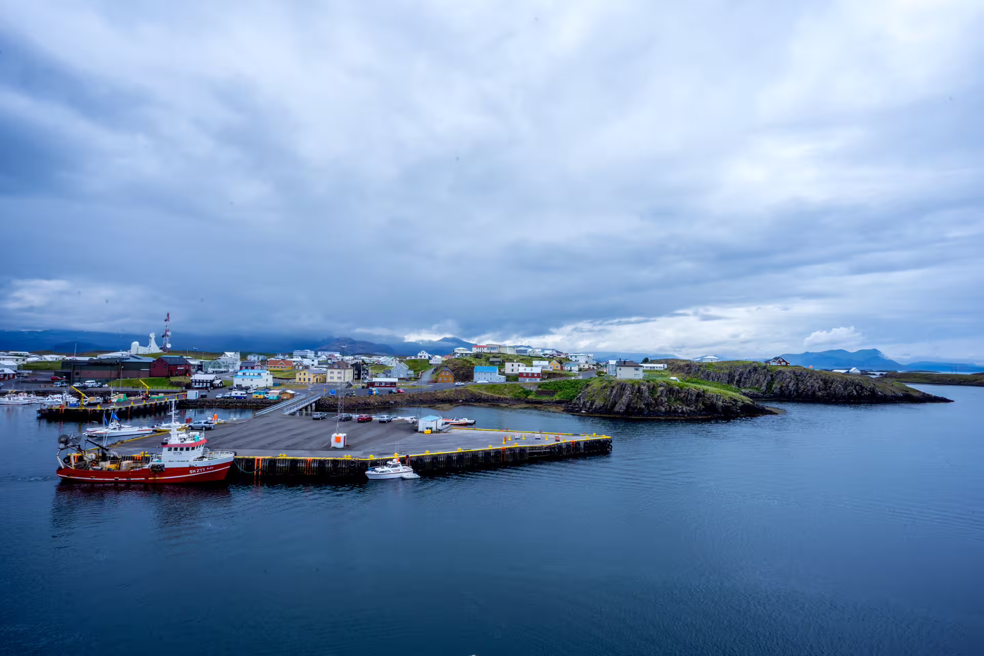 Red fishing boat docked at Stykkishólmur harbor with colorful town buildings and rocky coastline on Snaefellsnes Peninsula