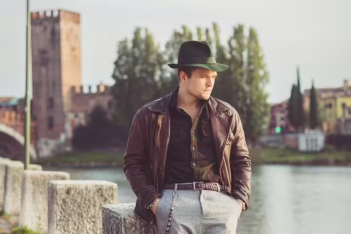 Stylish man in a hat and leather jacket posing by the river in Verona, showcasing a timeless, fashionable photoshoot.