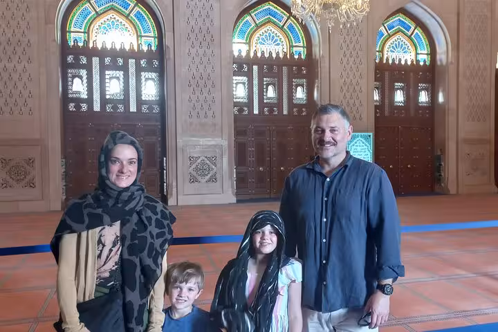 Family of tourists posing inside Sultan Qaboos Grand Mosque Muscat with stained glass windows and grand wooden doors