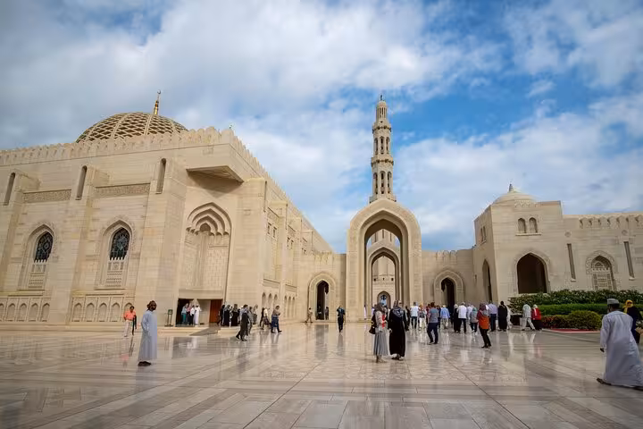 Visitors exploring the grand exterior courtyard and minaret of Sultan Qaboos Grand Mosque on a guided tour in Muscat