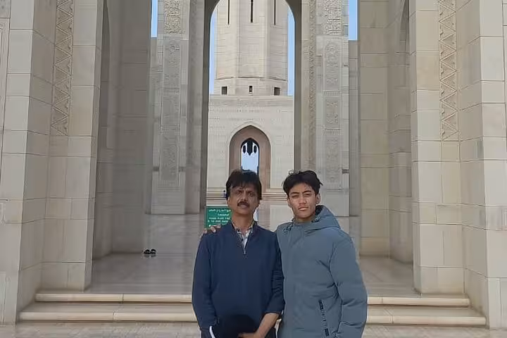 Two visitors posing under the grand arched entrance of Sultan Qaboos Grand Mosque on a guided sightseeing tour in Muscat