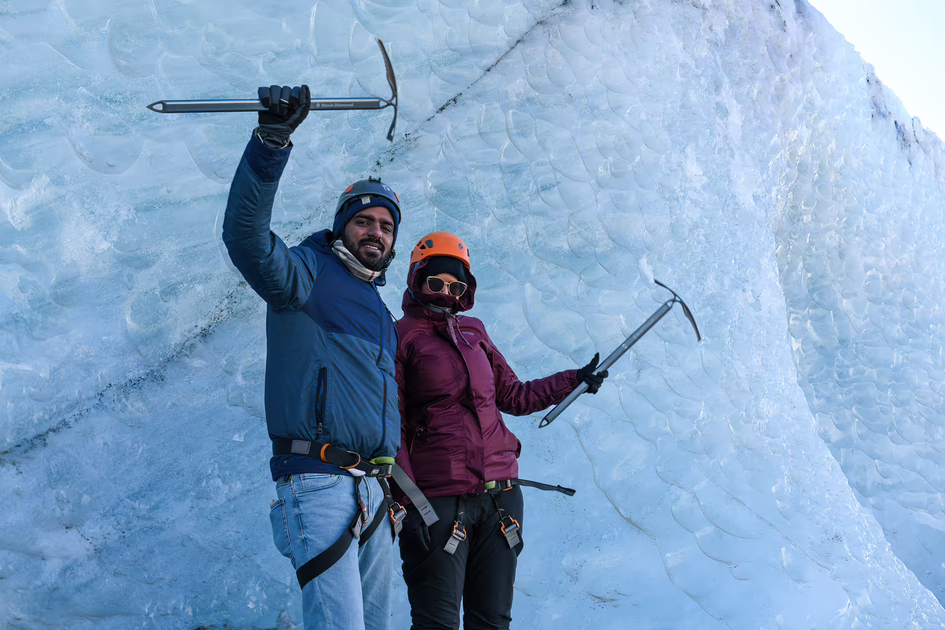 Two adventurers in helmets and harnesses hold ice axes beside a towering glacier wall on a summer highland tour.