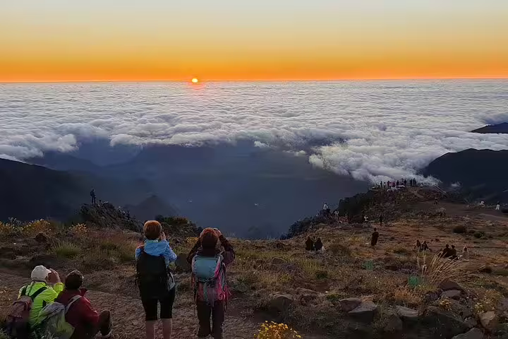 Tourists admire a breathtaking sunrise over a sea of clouds at Areeiro Peak, Madeira, offering stunning views and photo opportunities.