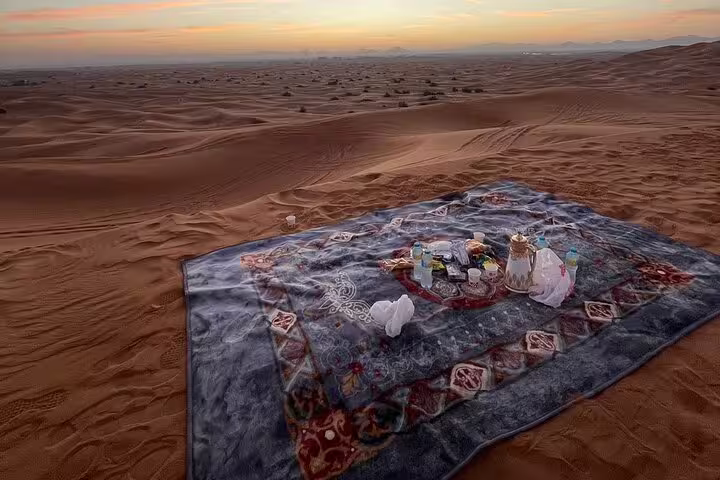 Luxurious picnic setup on a patterned rug at sunrise in the Dubai desert, offering a serene refreshment spot.
