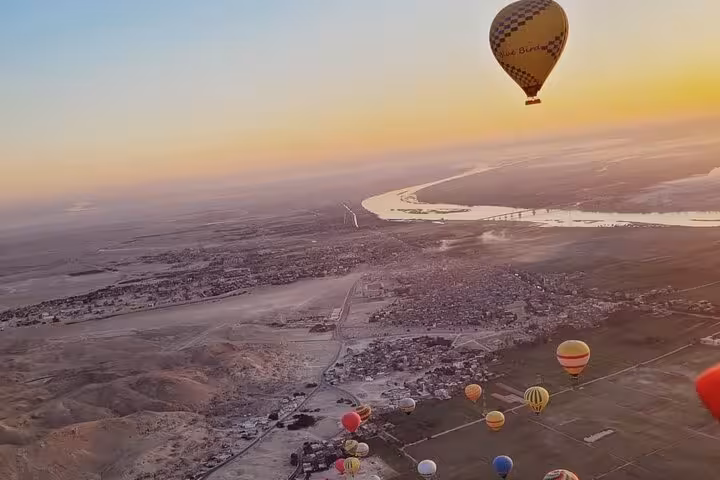 Hot air balloons floating over Luxor West Bank at sunrise, with a panoramic view of the Nile River and desert landscape.