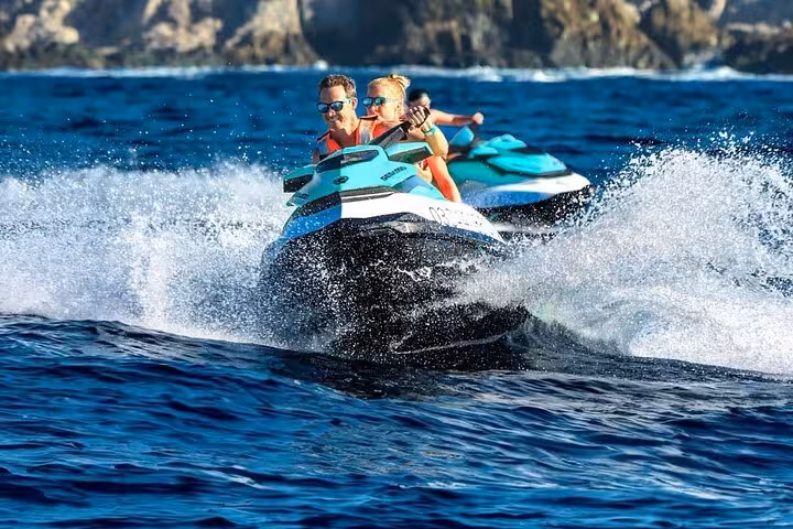 Two people riding teal Sea-Doo jet skis at speed on blue Mediterranean waters near rocky coastline