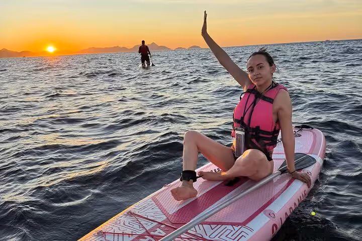 Join a sunrise SUP adventure in Copacabana with a woman waving from her board as the sun rises over calm waters.