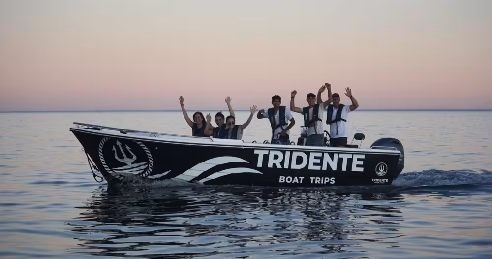 Excited guests wave aboard a Tridente Boat Trips vessel during a stunning sunset Benagil Cave tour along the Algarve coastline.