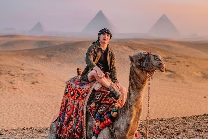 Traveler riding a camel at sunset with Giza Pyramids in the distance, part of camel ride and Sound & Light show