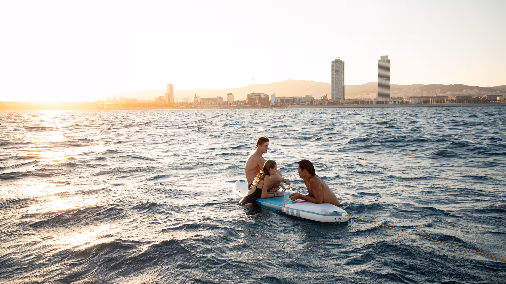 Guests relaxing on a paddleboard beside the catamaran at sunset with Barcelona skyline in the distance