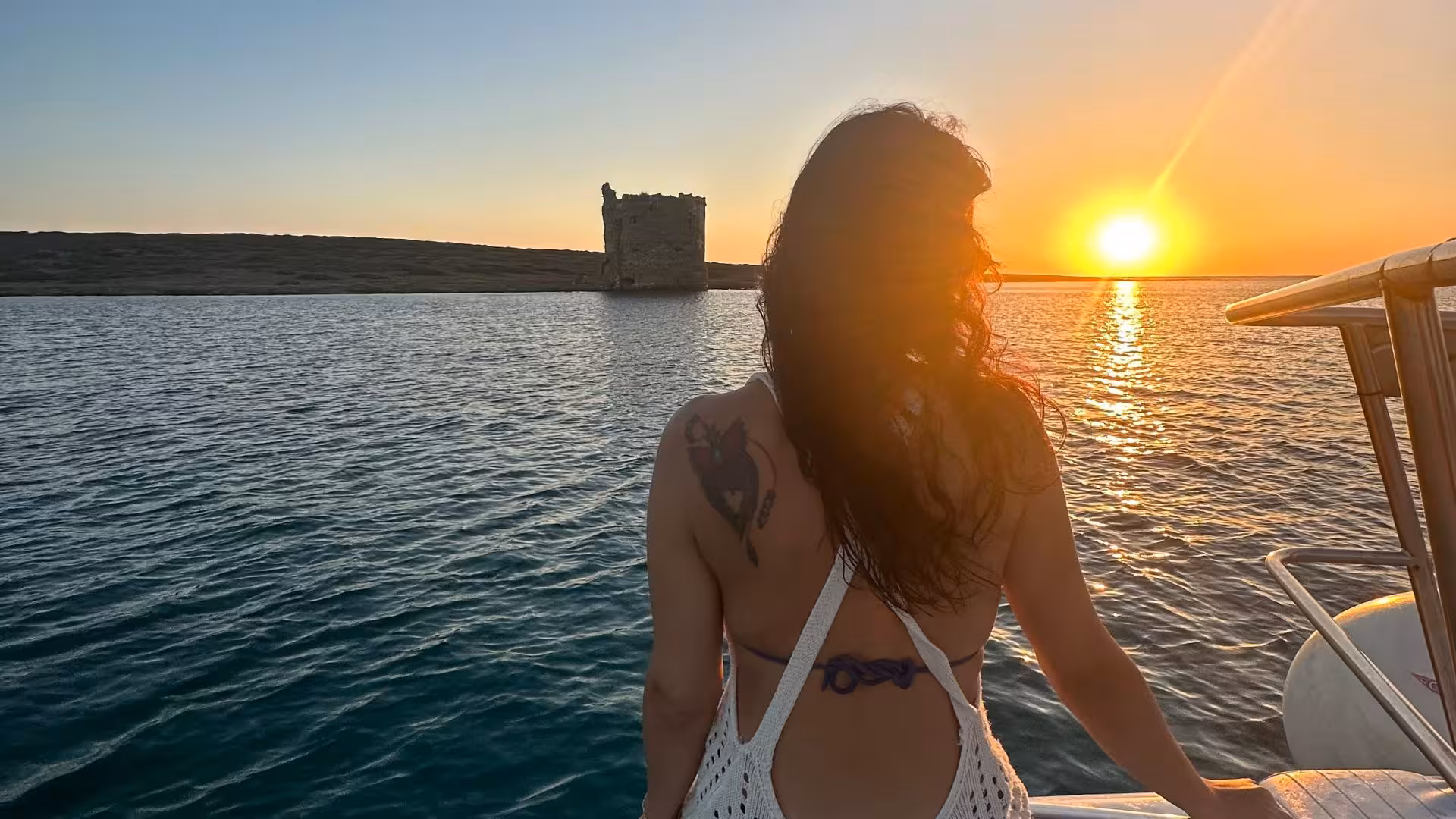 Woman enjoying a sunset catamaran tour in the Gulf of Asinara with a view of a historic tower and tranquil waters.