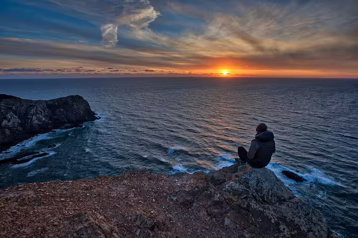 Person sits on cliff edge at sunset overlooking ocean during Algarve Natural Park West Coast tour.