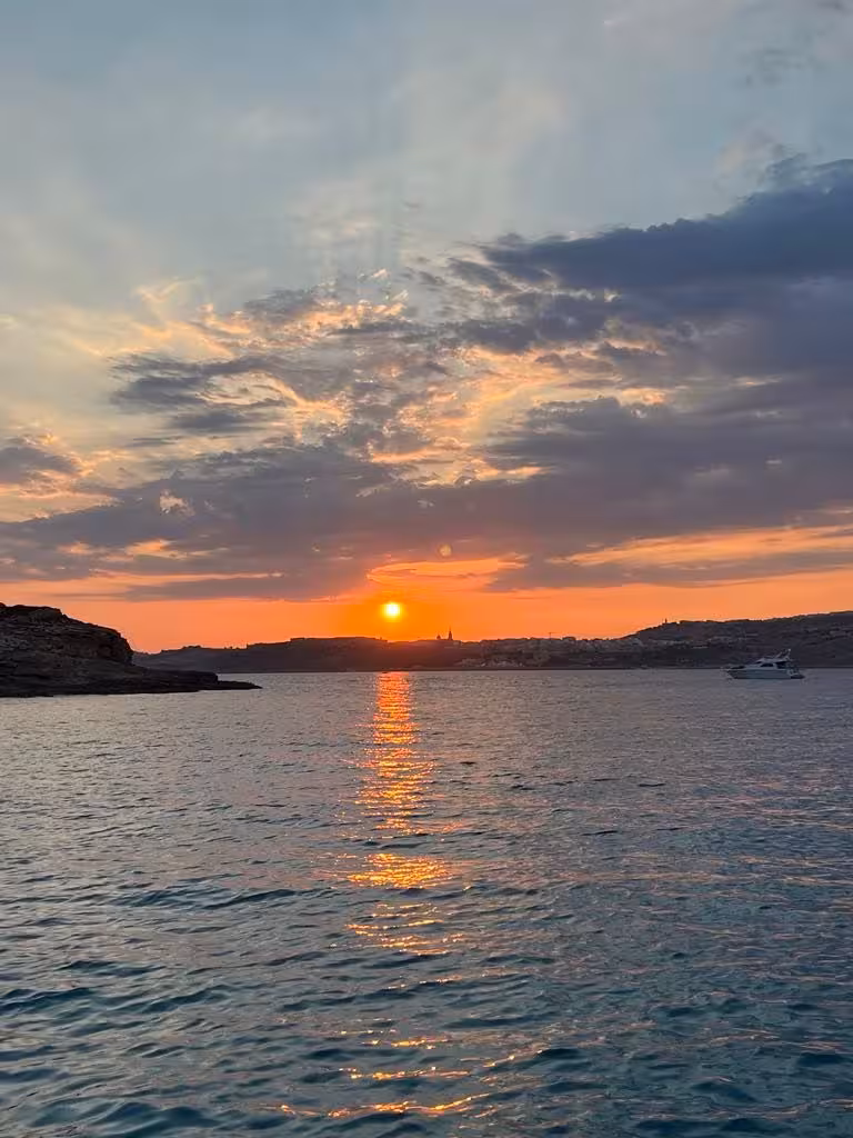 Sunset over Comino waters on Blue and Crystal Lagoons cruise, golden sky reflections and boat near Malta coast