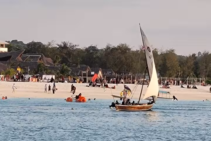 Dhow sailing near Nungwi beach at sunset with tourists enjoying Zanzibar's tranquil blue waters.