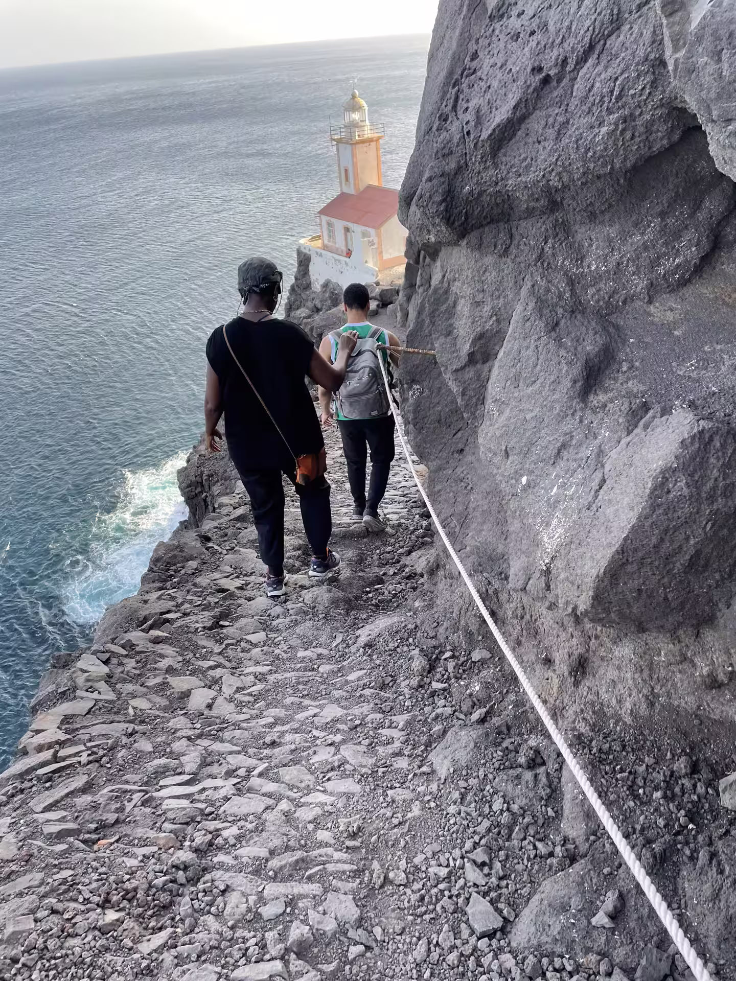 Hikers traverse rocky coastal path toward Lighthouse Dona Amélia at sunset, offering stunning ocean views and adventure.