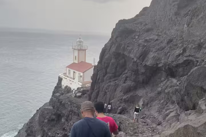 Hikers navigate rocky cliffs towards the historic Lighthouse Dona Amélia, overlooking the ocean during a scenic sunset tour.