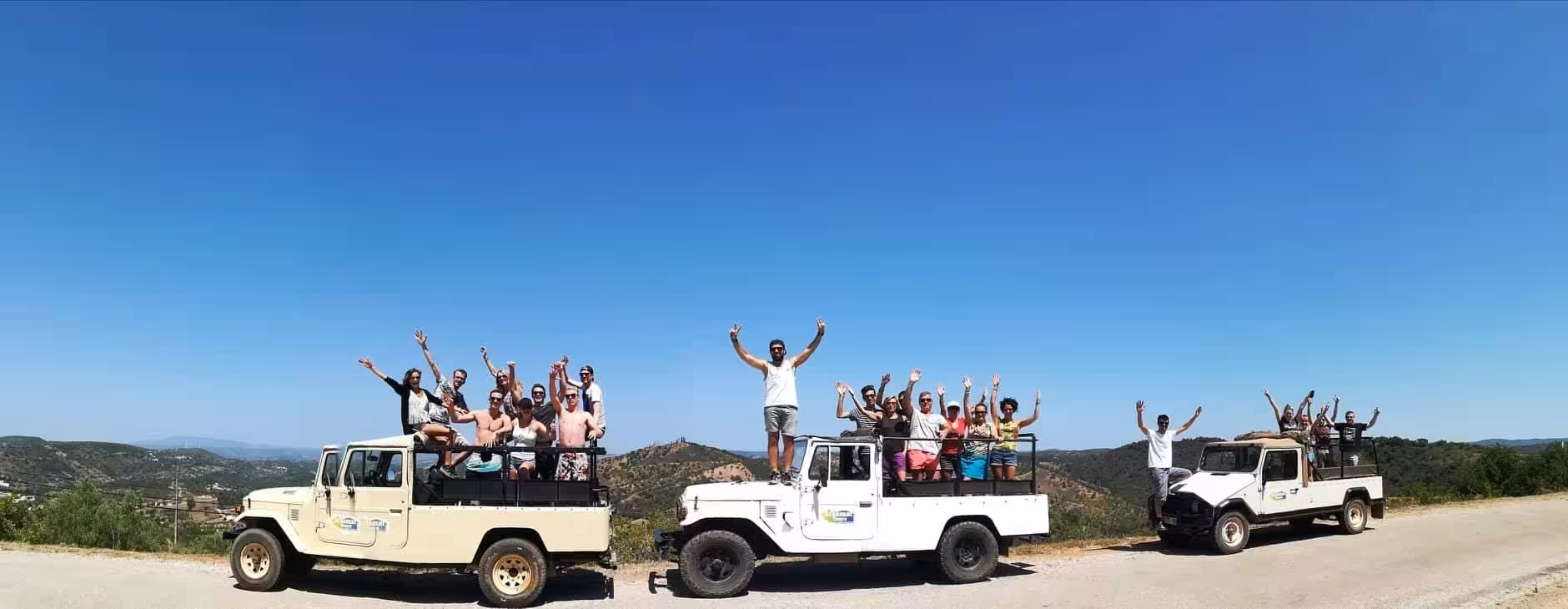Open-top jeeps full of excited travelers on an Algarve sunset safari tour, parked on a scenic hill road under clear blue sky.