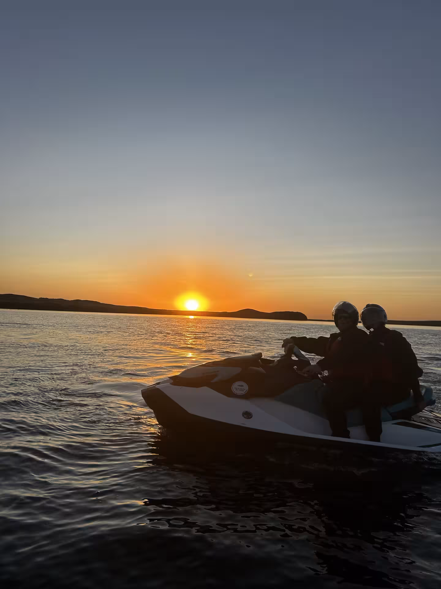 Two riders on a jet ski silhouetted against a golden sunset over the calm waters of Reykjavik Bay Iceland.