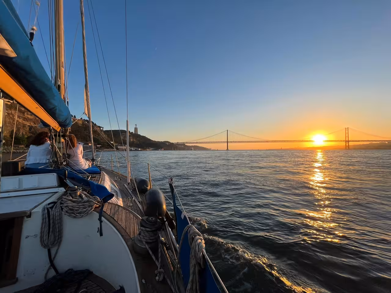 Shared sunset sailboat tour gliding under Lisbon’s 25 de Abril Bridge with golden Tagus River views and scenic coastline