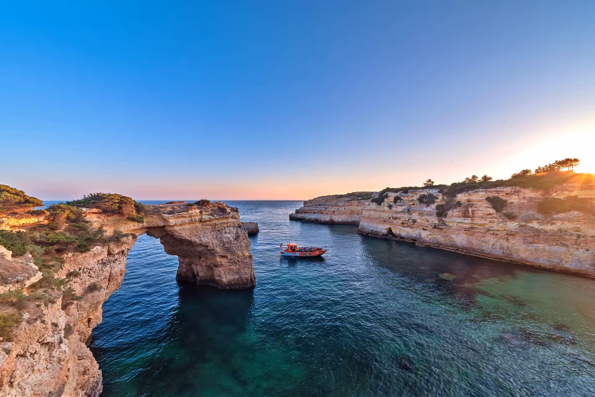 Sunset tour boat cruising between dramatic Algarve sea cliffs and natural rock arch over crystal-clear Atlantic waters