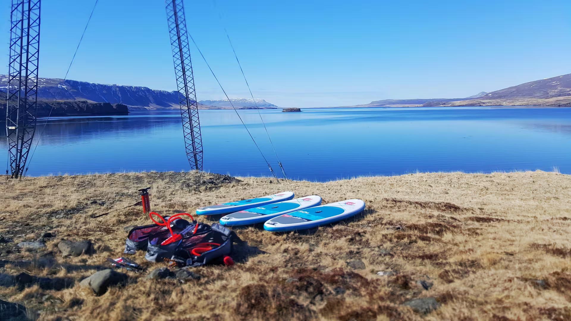Three paddle boards and gear on a grassy shoreline overlooking the serene, blue waters of the Forgotten Fjords under clear skies.