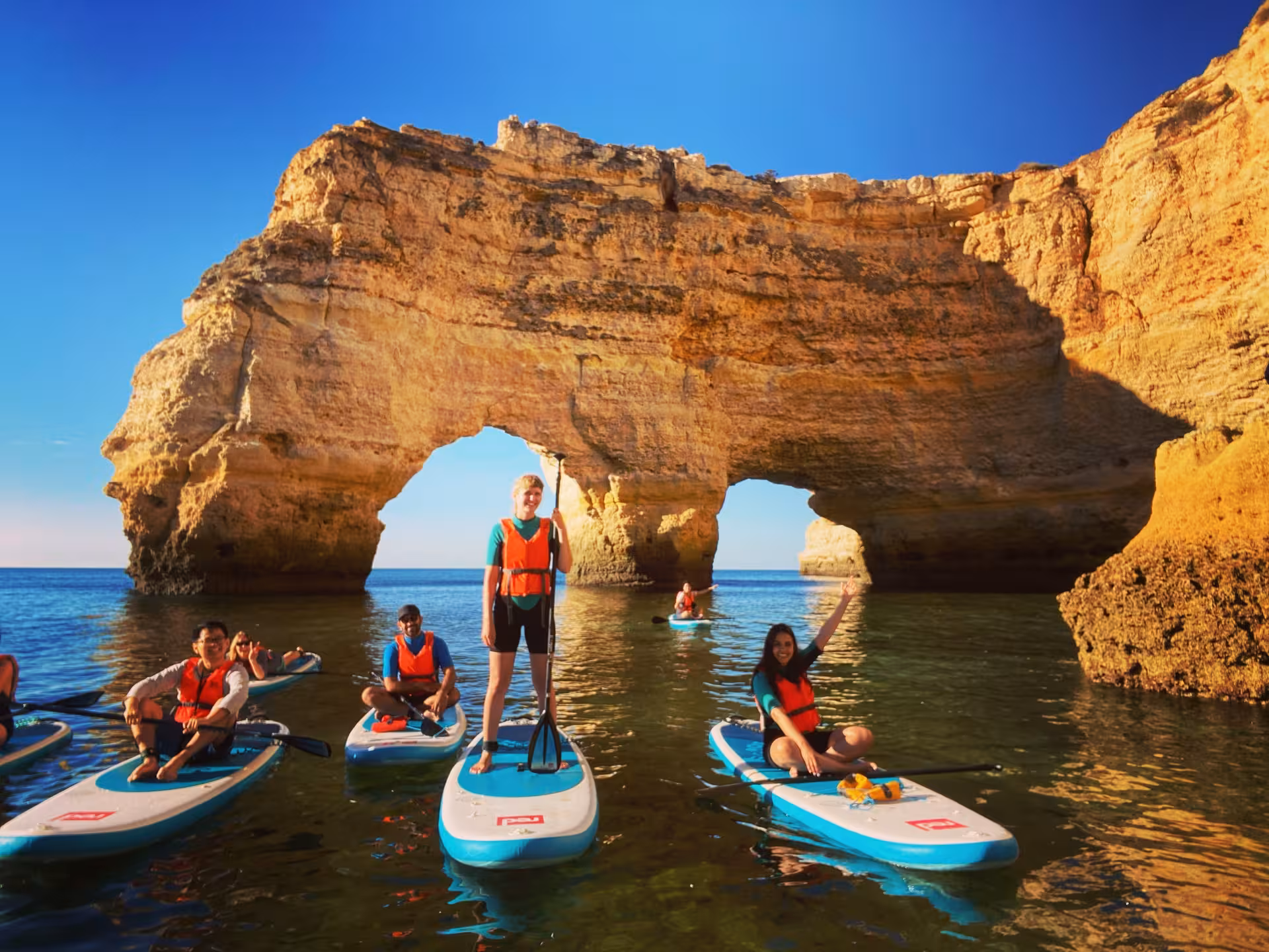 Group of friends stand up paddleboarding by golden sea caves and rock arches on a sunny Algarve coast SUP rental tour