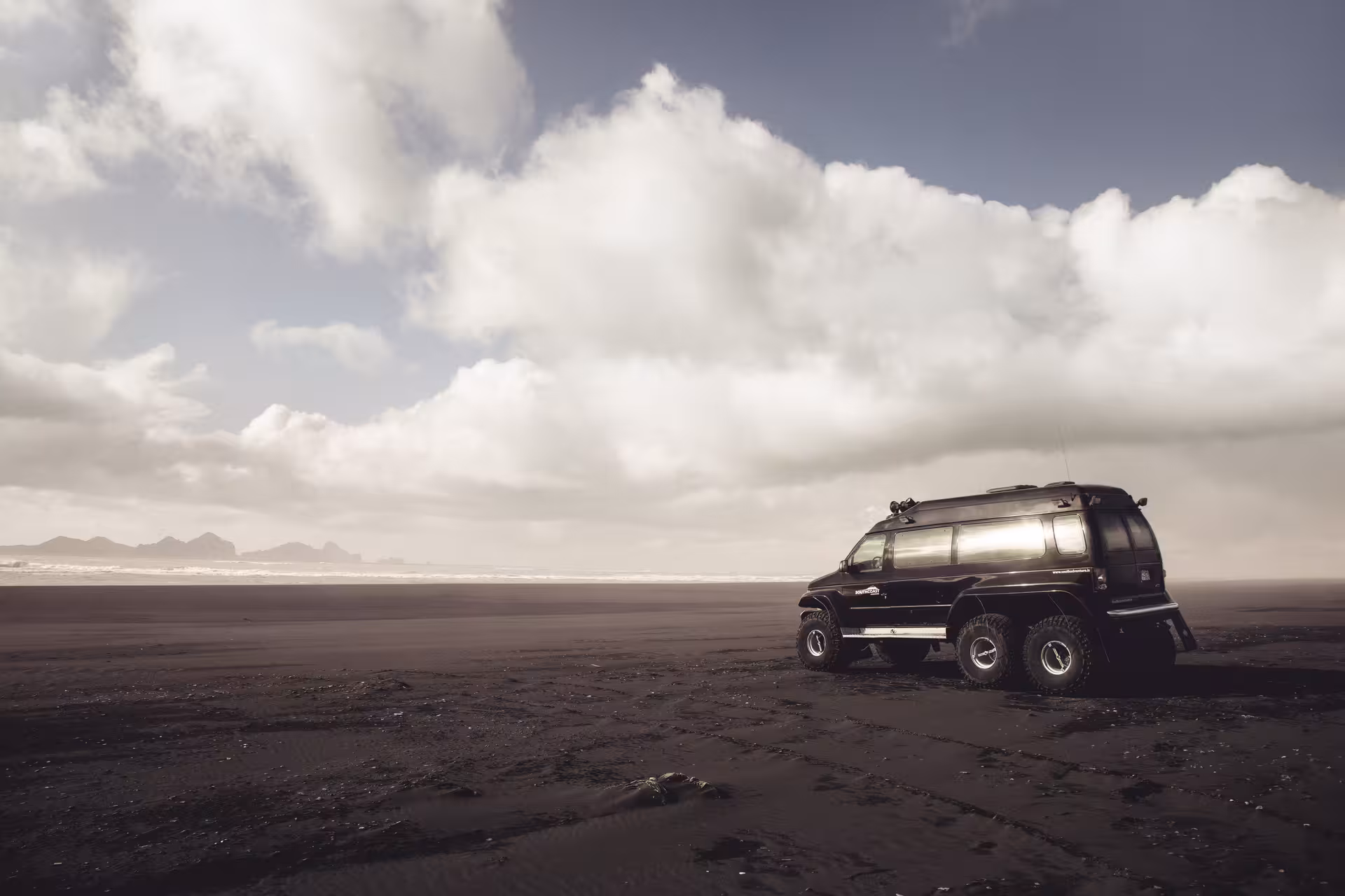 Black 6-wheel super jeep on Iceland's dark volcanic black sand beach with dramatic cloudy sky