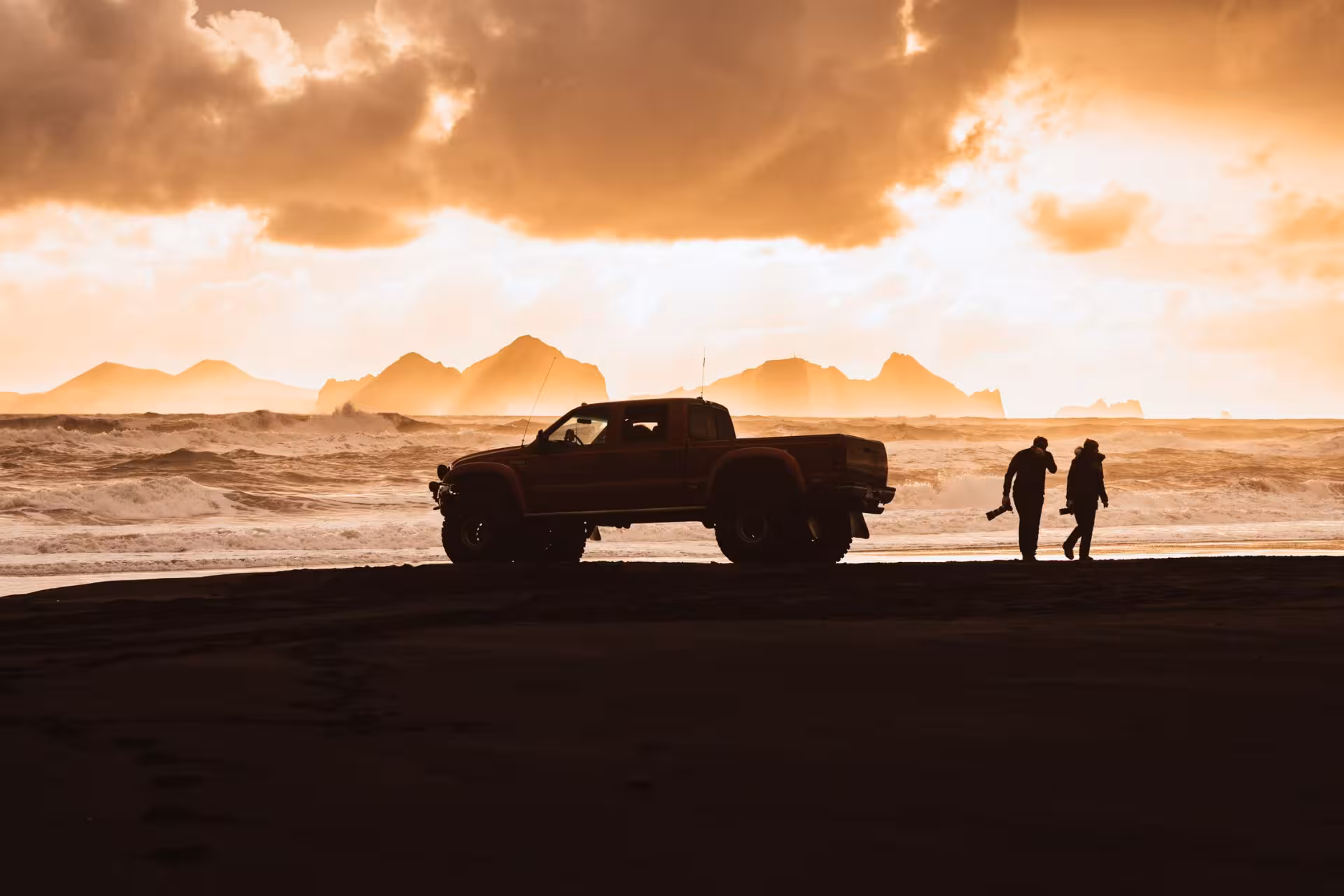 Super jeep and two photographers silhouetted on black sand beach with dramatic sunset and mountains near Thorsmork