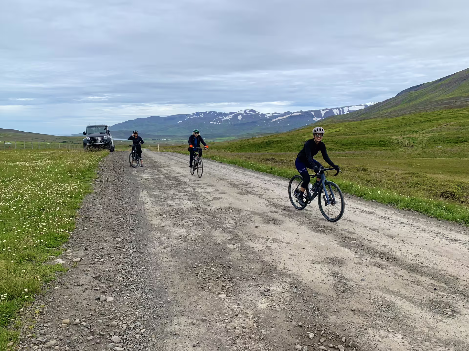 Three cyclists riding on a gravel road through green Icelandic highlands with snow-capped mountains and a Super Jeep behind them.