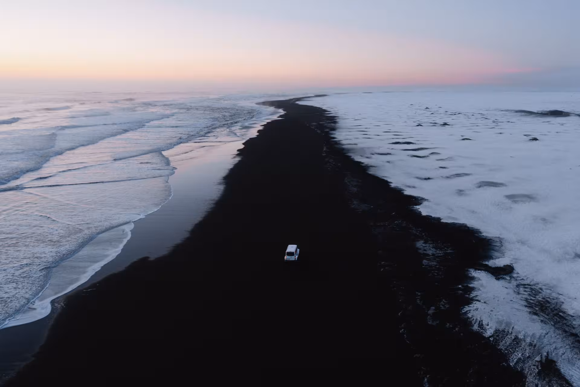 Aerial view of a white super jeep driving along Iceland's black sand beach at sunset in winter