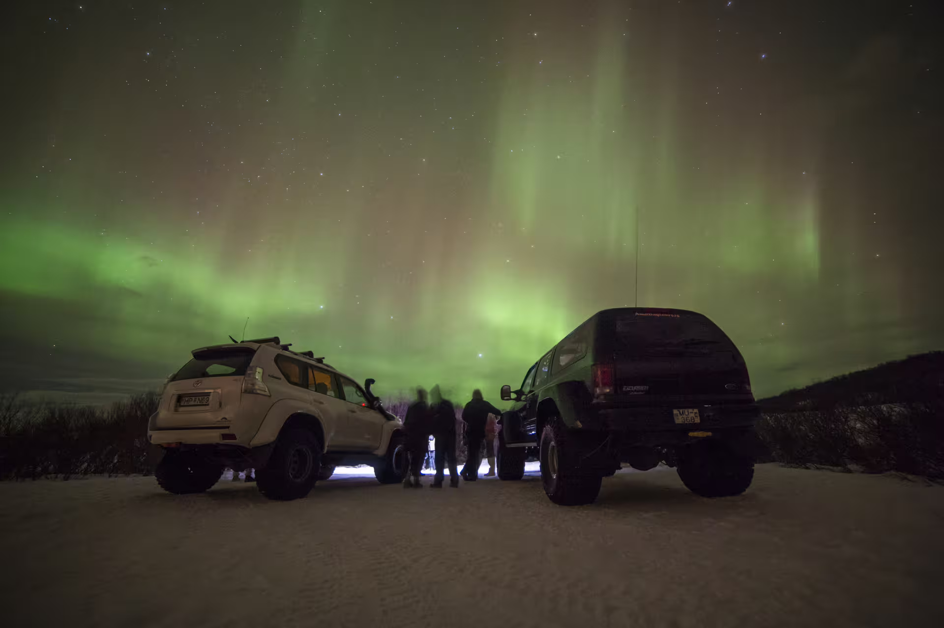 Two super jeeps parked under vivid green northern lights aurora in snowy Iceland at night with silhouetted visitors