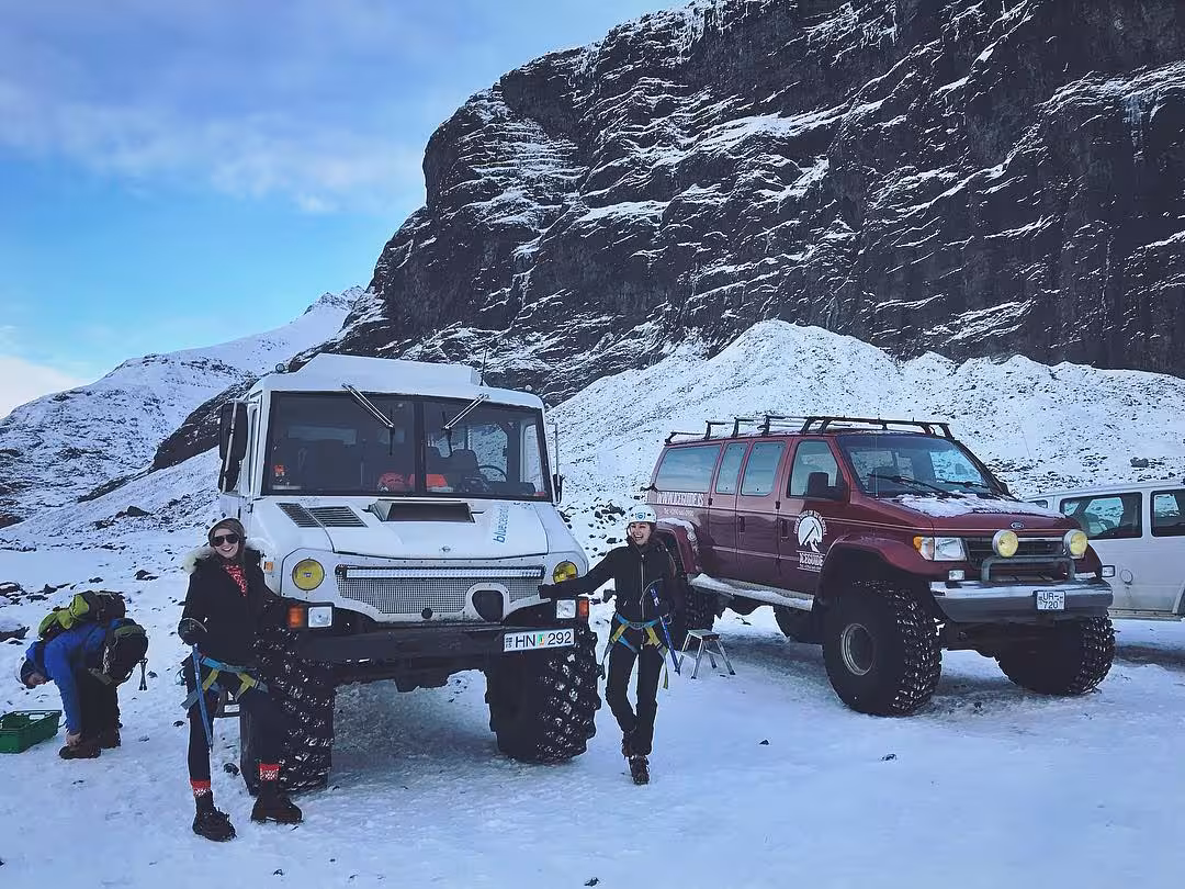 Two people posing beside a white Snowcat and red super jeep on snow in front of a rocky mountain in Iceland