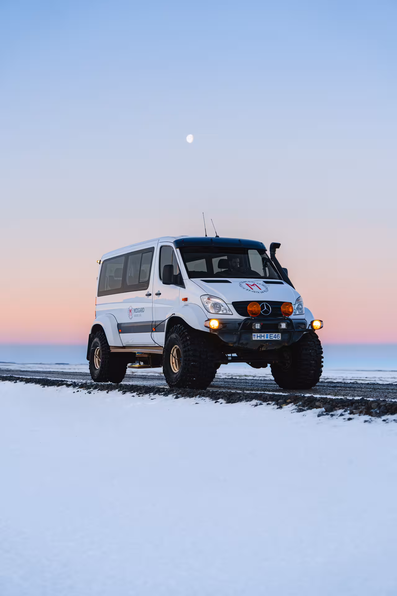 White lifted Mercedes Sprinter Super Jeep on snowy landscape at sunset with moon visible in sky