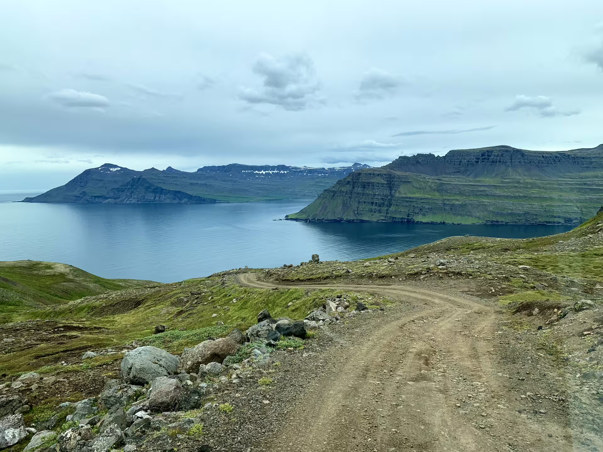 Winding gravel mountain road overlooking a calm fjord with dramatic cliffs and green hillsides in Iceland