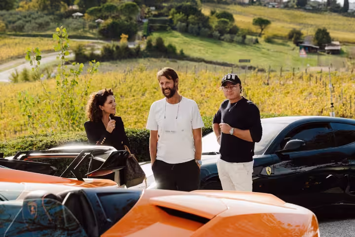 Three people smiling beside supercars with Chianti vineyard hills in background, Siena Italy