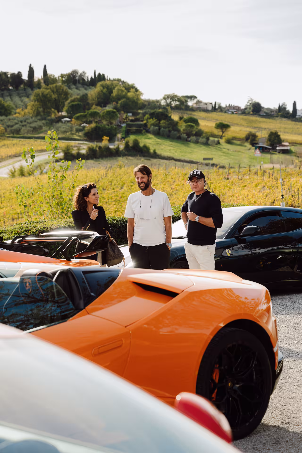 Three people chatting beside Lamborghini supercars in a Tuscan vineyard landscape