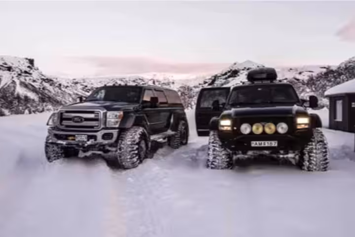 Two black lifted superjeeps driving through deep snow with snowy mountains in background