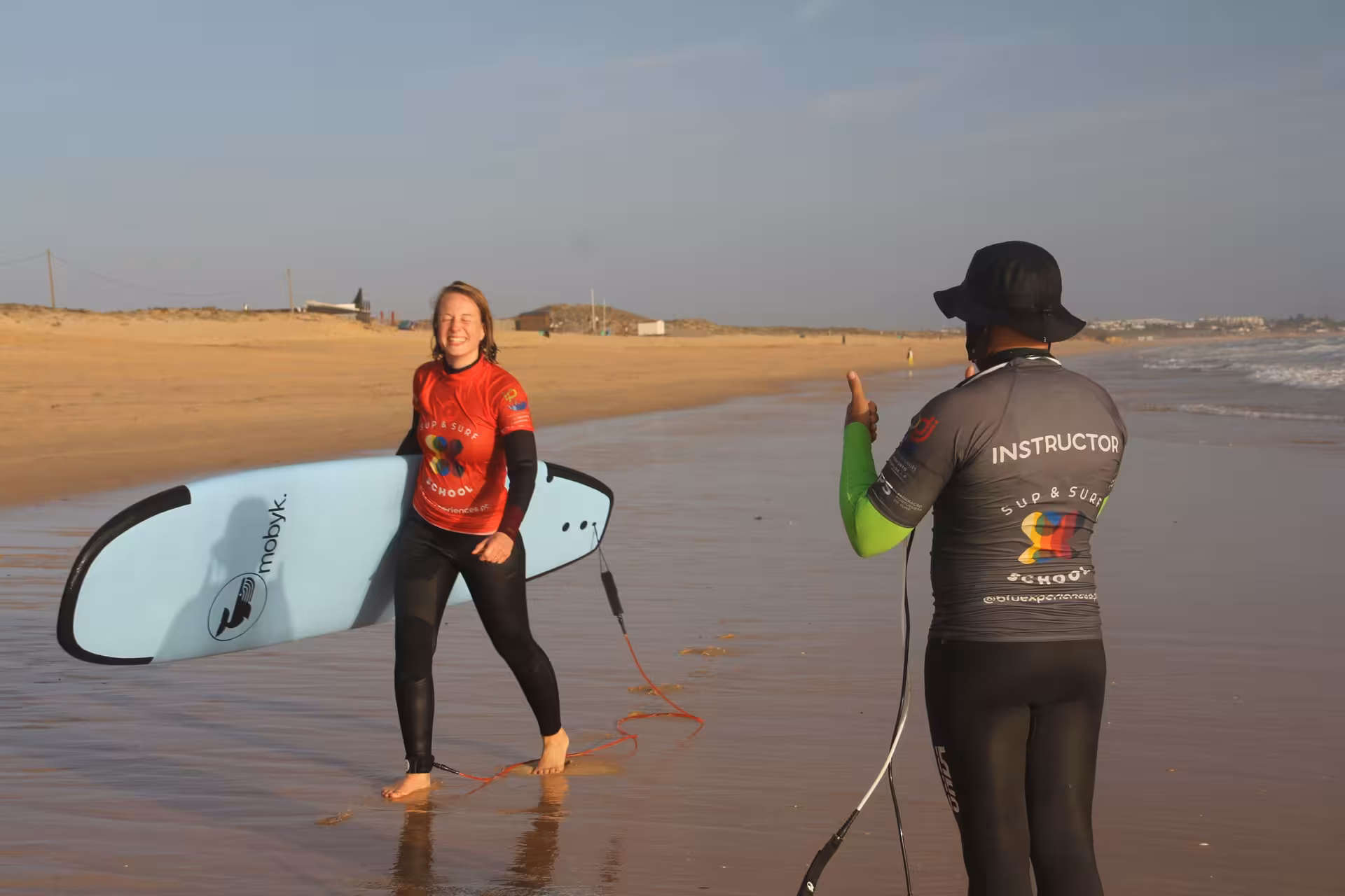 Surf student carrying a blue foam board and chatting with instructor on a wide sandy beach after group lesson