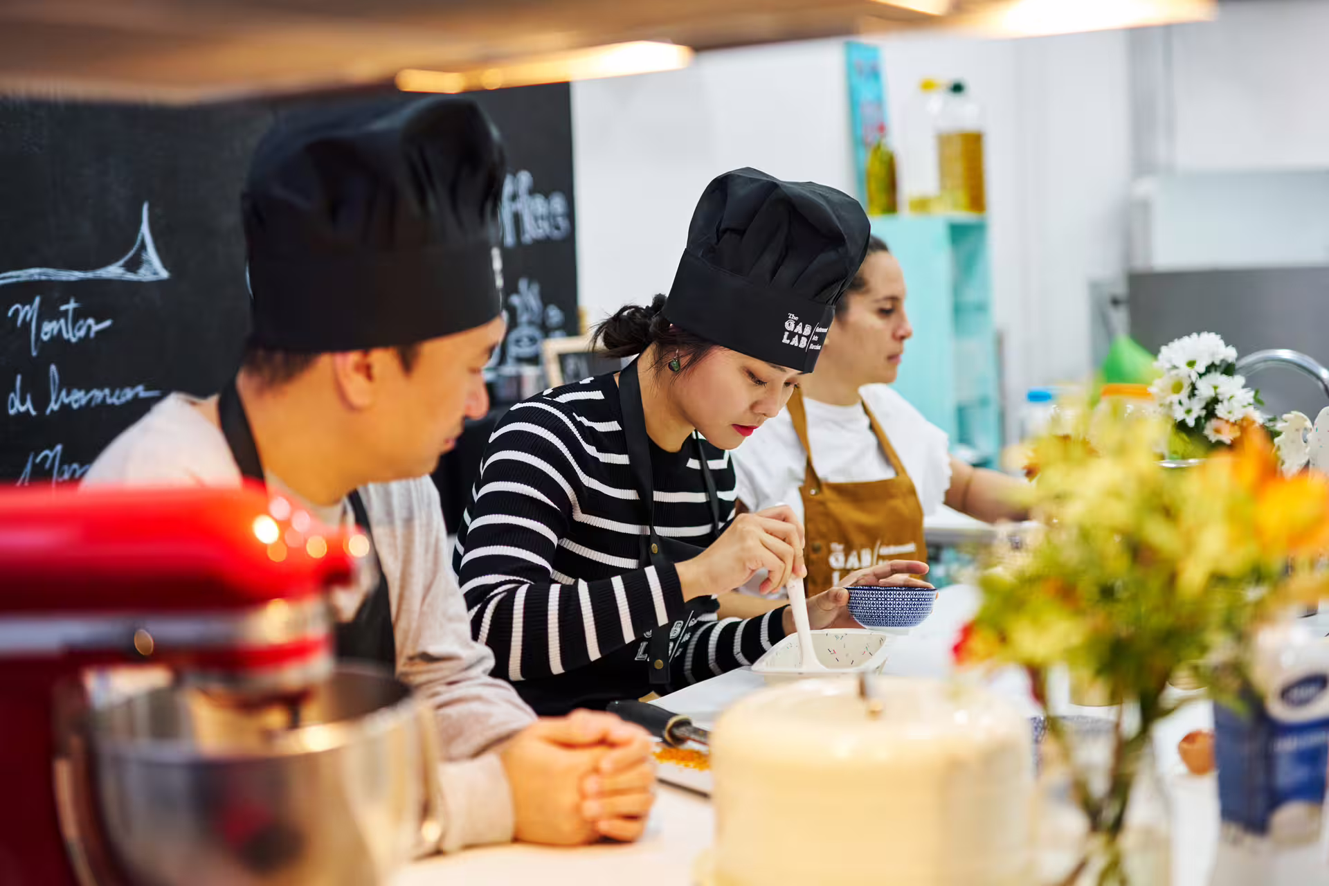 Participants in chef hats engage in hands-on baking at the Sweet Escape: Bake & Take Workshop, surrounded by vibrant kitchen decor.