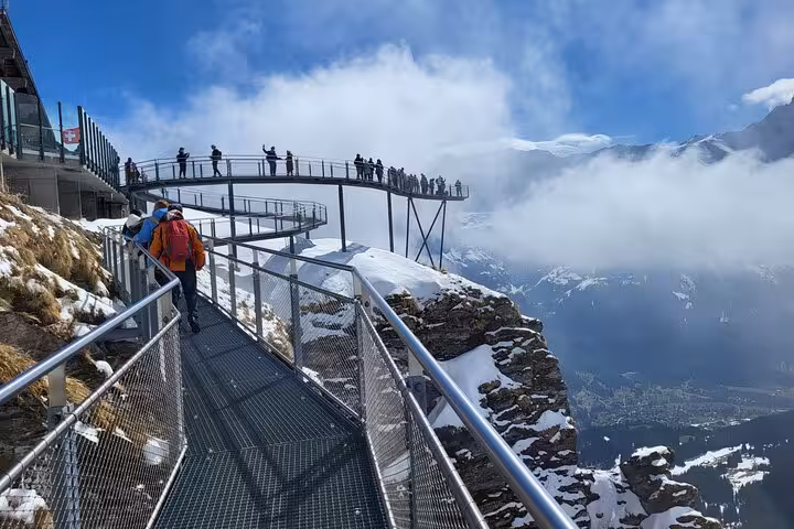 Tourists enjoying panoramic views from a scenic walkway in the snow-capped Swiss Alps on a clear day.