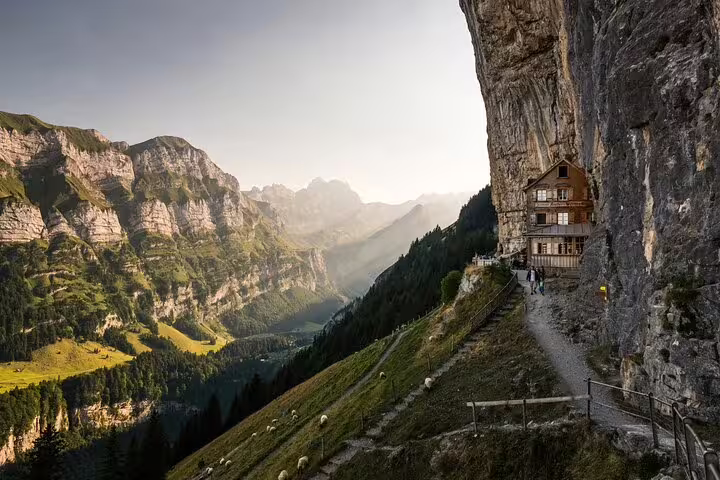 Wooden alpine guesthouse built into steep cliff face overlooking lush Swiss mountain valley at golden hour