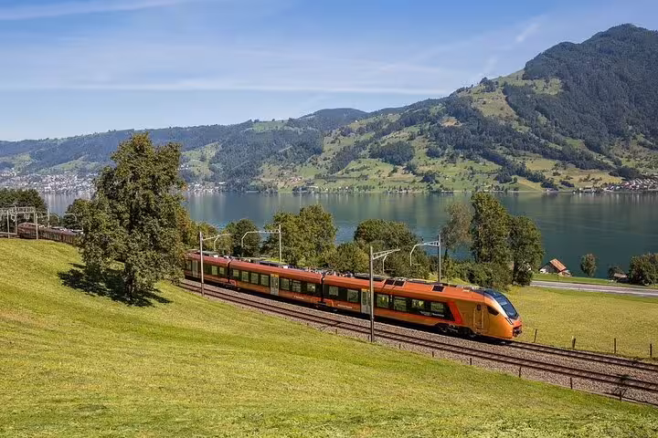 Orange Swiss train journeying beside a tranquil lake with picturesque mountain views and verdant landscapes.