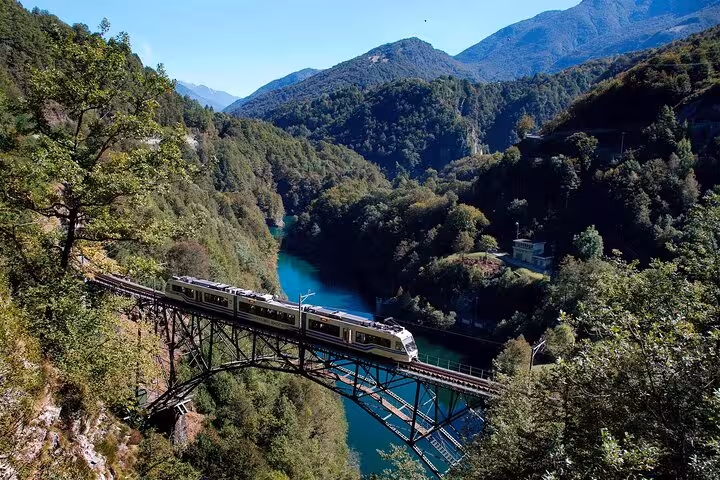 Swiss train crosses a high bridge over a turquoise river, surrounded by scenic mountains and dense forest.