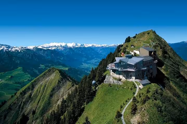 Stunning panoramic view from a Swiss mountain peak, featuring a modern summit building and distant snow-capped Alps.