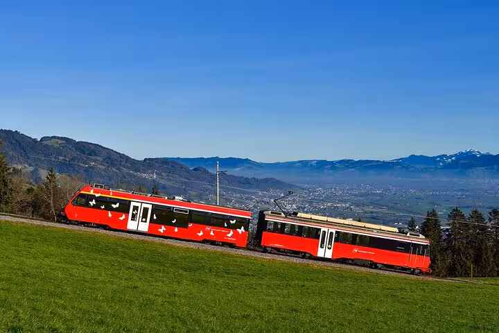 Scenic Swiss train with vibrant red carriages traveling through lush countryside and mountain backdrop on a clear day.