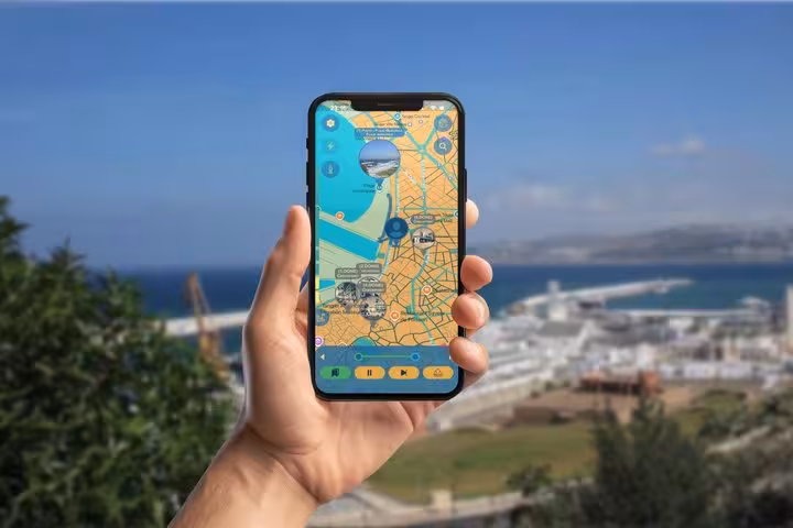 Hand holding smartphone displaying GPS walking tour map of Tangier International Zone with coastal city view