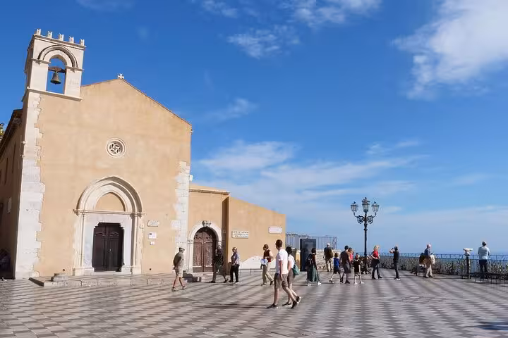 Visitors enjoy the scenic view from Taormina's piazza, featuring a historic church and panoramic sea vistas.